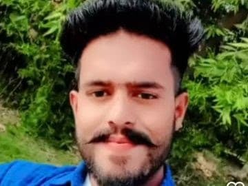 A Hindu man with a mustache and beard, smiling, outdoors with green plants in the background.