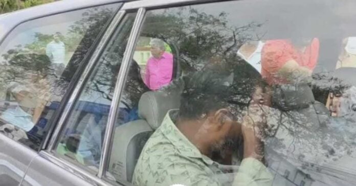 Man using mobile phone while seated in a car, with reflections of trees and people outside.