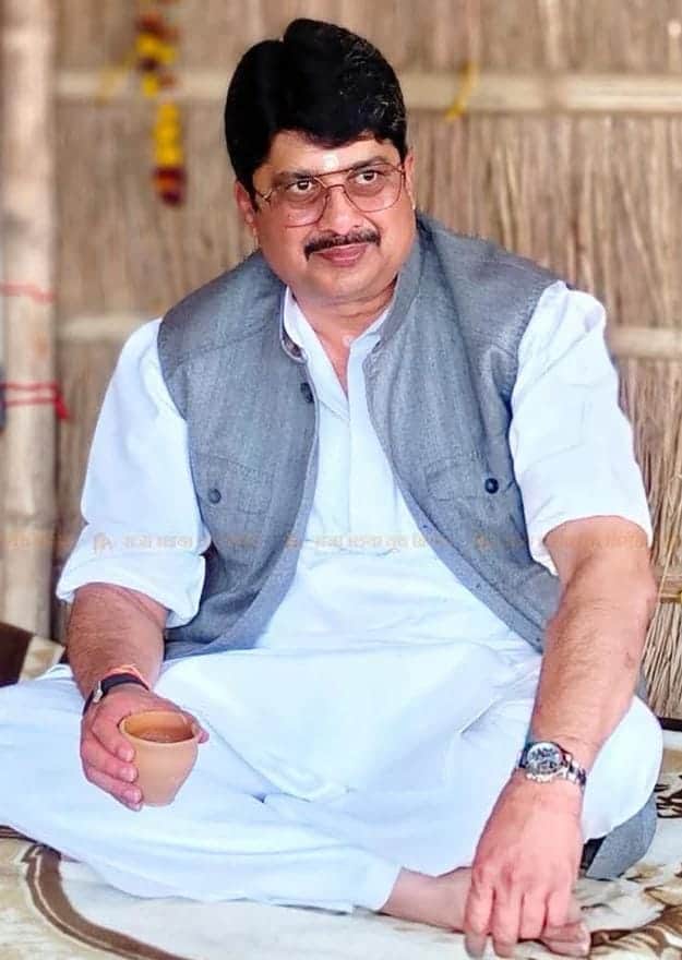 Hindu man in traditional attire sitting cross-legged, holding a cup, in a cultural setting.