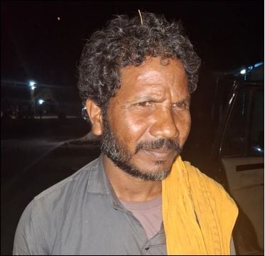 Man with curly hair and beard, wearing a yellow cloth over shoulder, nighttime background.