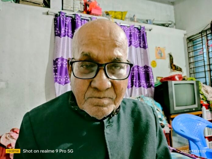 Portrait of Rasendra Datta Chowdhury, an elderly man with glasses, in a room with colorful curtains.