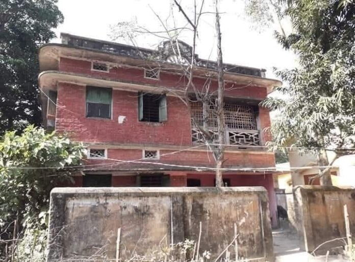 Dilapidated brick house with overgrown trees and weathered wall in urban setting.