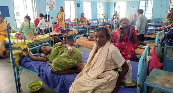 A woman lying on hospital bed with other patients and staff in West Bengal healthcare facility.