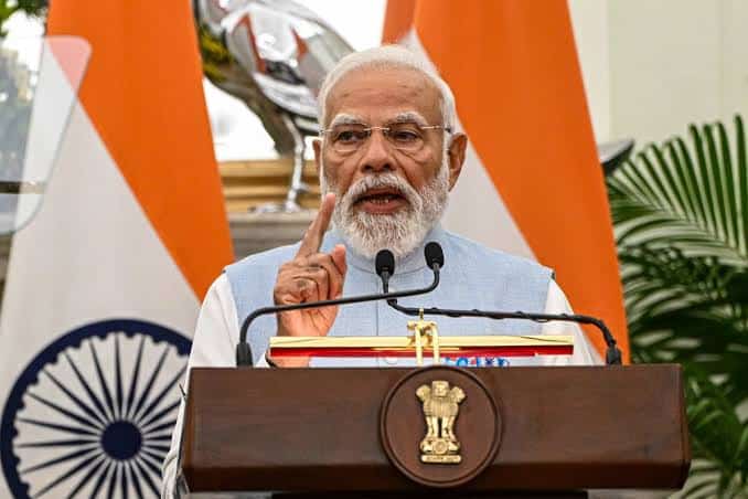 Indian Prime Minister Narendra Modi delivering a speech at a public event with Indian flags in the b.